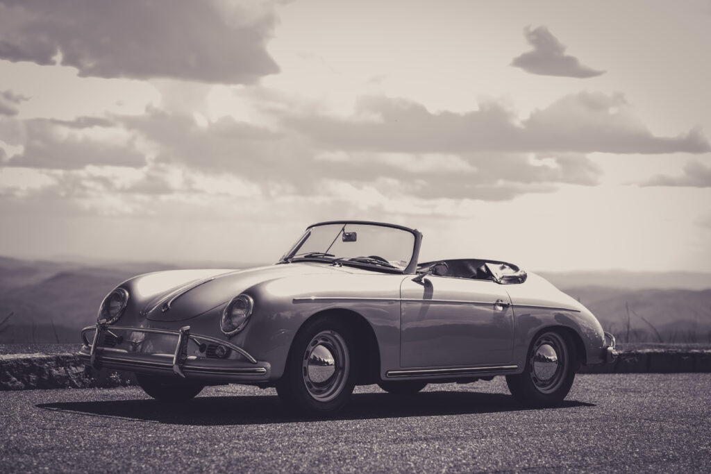 Zwart-witbeeld van een klassieke cabriolet auto op een asfaltweg met heuvels en een wolkenlucht op de achtergrond.