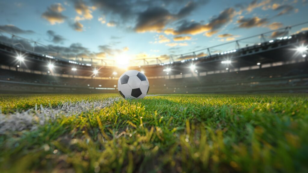 Close-up van een voetbal op groen gras in een groot voetbalstadion met felle lichten en een bewolkte avondlucht.